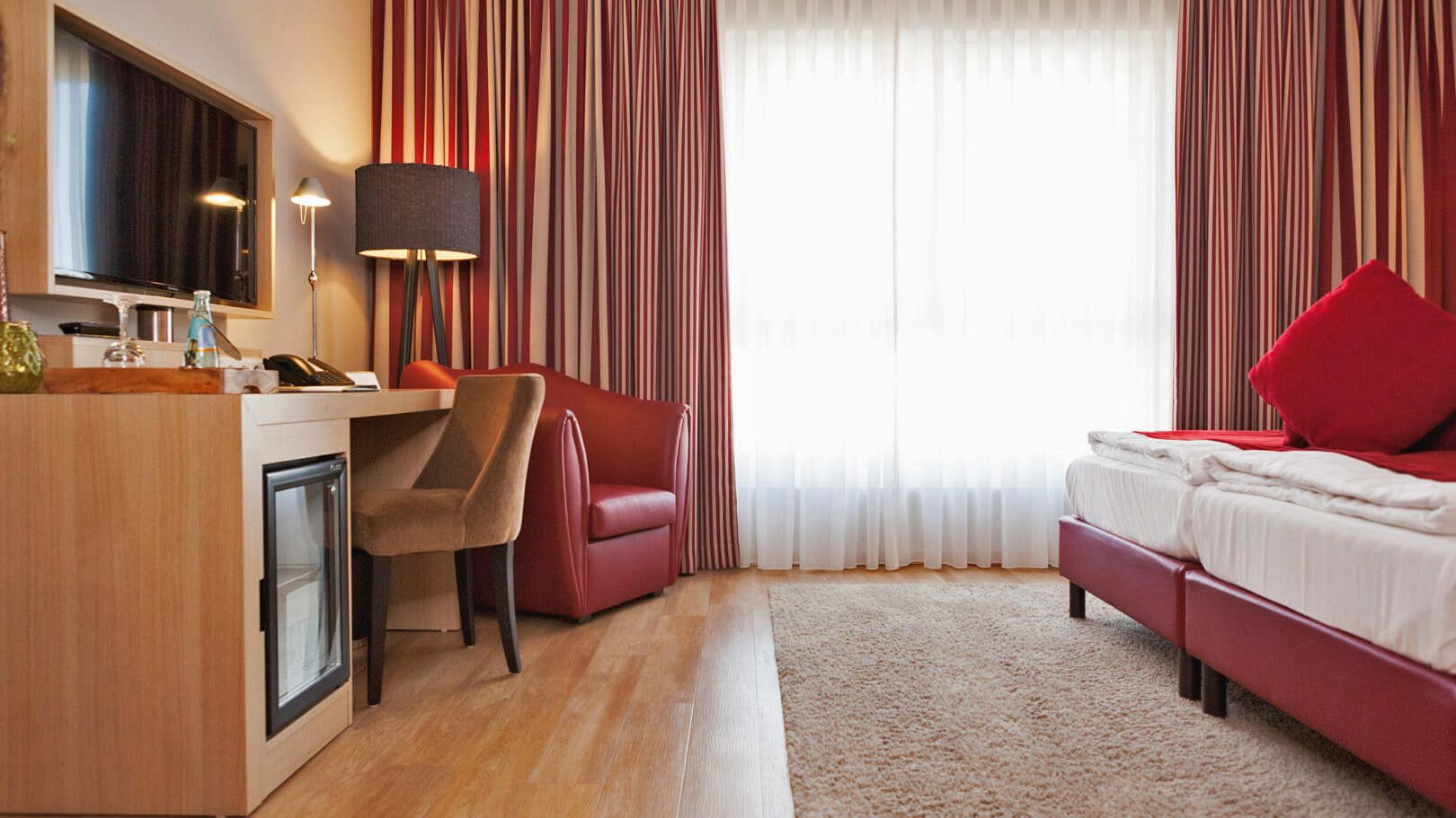 Zimmer der Kategorie Spaetlese im Weinhotel Kaisergarten Alzey Blick zum Schreibtisch, Stuhl TV und Minibar Sessel aus rotem Leder sowie auf das geraeumige Doppelbett