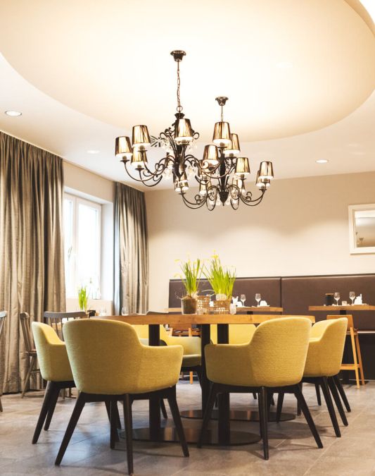 A view of the breakfast room at the Weinhotel Kaisergarten, seen from the buffet area, featuring wooden tables, several armchairs, and two designer chandeliers